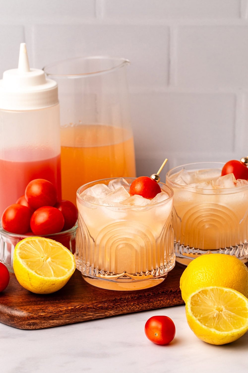 sparkling tomato lemonade in a glass with ice garnished with a cherry tomato. The glass is on a wood cutting board in a white kitchen